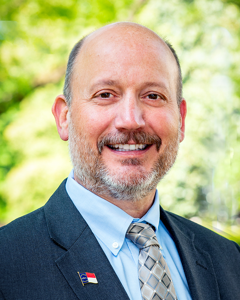 A smiling middle-aged man with dark eyes, hair on the sides of his head but bald on top, salt and pepper hair, moustache, and beard, dark gray suit, light blue shirt, light and dark silver tie with alternating diamond patterns, and a North Carolina flag pin on his suit jacket.