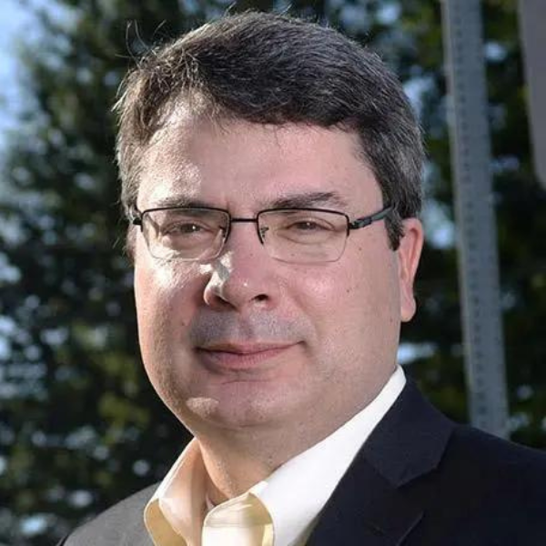 A straight-faced middle aged white man with a black jacket, white shirt, and gold tie, wtih trees in the background