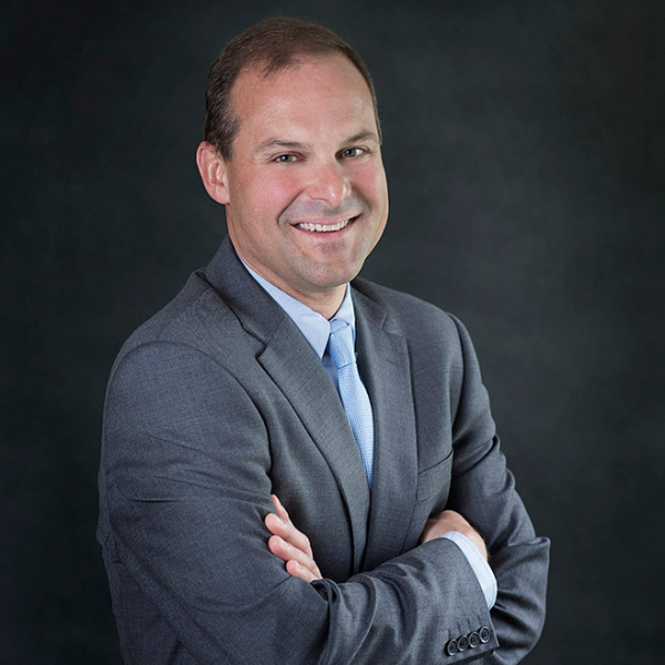 A smiling middle-aged man turned slightly to his right, brown hair, gray jacket, in the arms crossed business pose