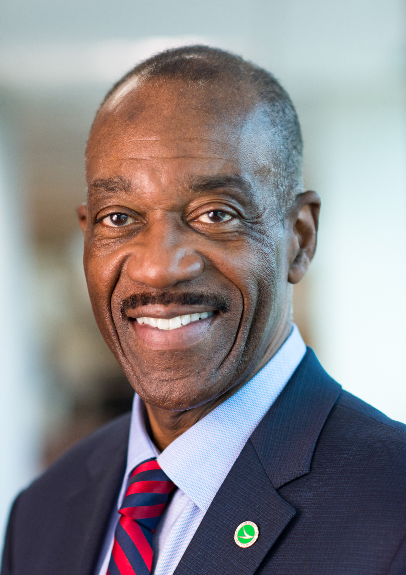 A smiling, older African American man with a moustache, navy suit jacket, light blue shirt, red and blue tie with alternating stripes, and a green pin with white markings