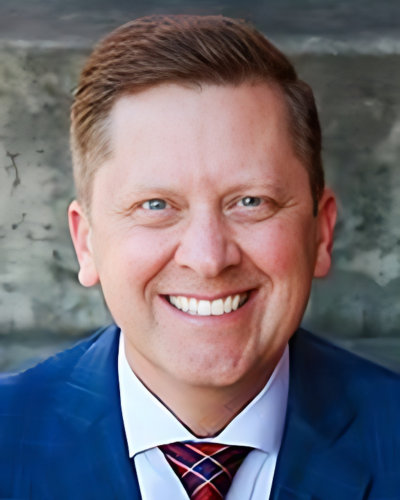 A smiling man with red hair and blue eyes, a plaid blue/light blue suit jacket, white shirt, and red and blue tie with gold and white accent lines