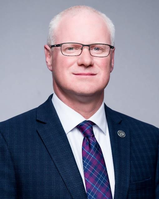 A balding man with white hair, glasses, a navy suit jacket, blue tie, and lapel pin