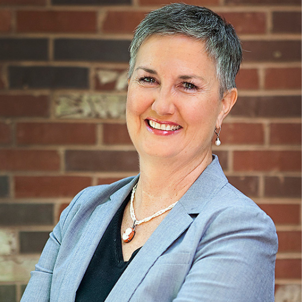 A smiling white woman with short salt and pepper hair, gold necklace with pendant, black V-neck shirt, and light blue jacket