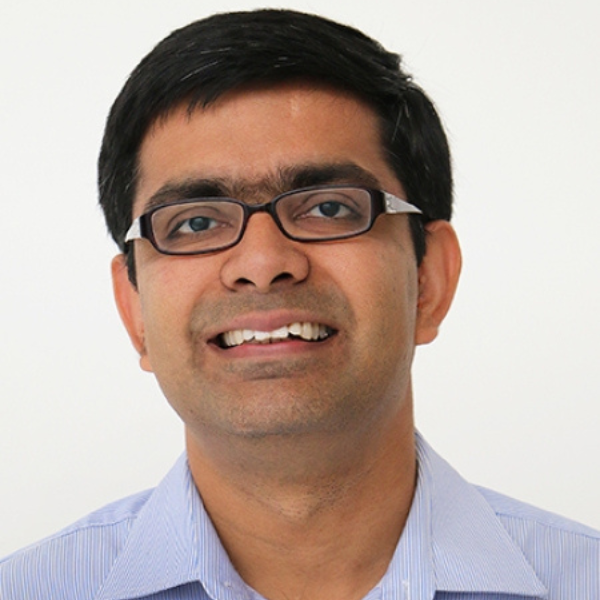 A smiling Indian man with a light blue shirt and black wire-rimmed glasses