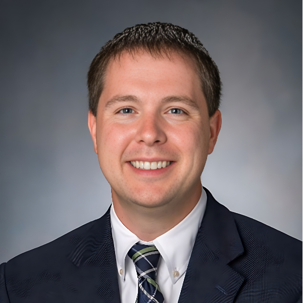 A smiling young man with short brown hair, a white shirt, black jacket and a blue, green, and white st