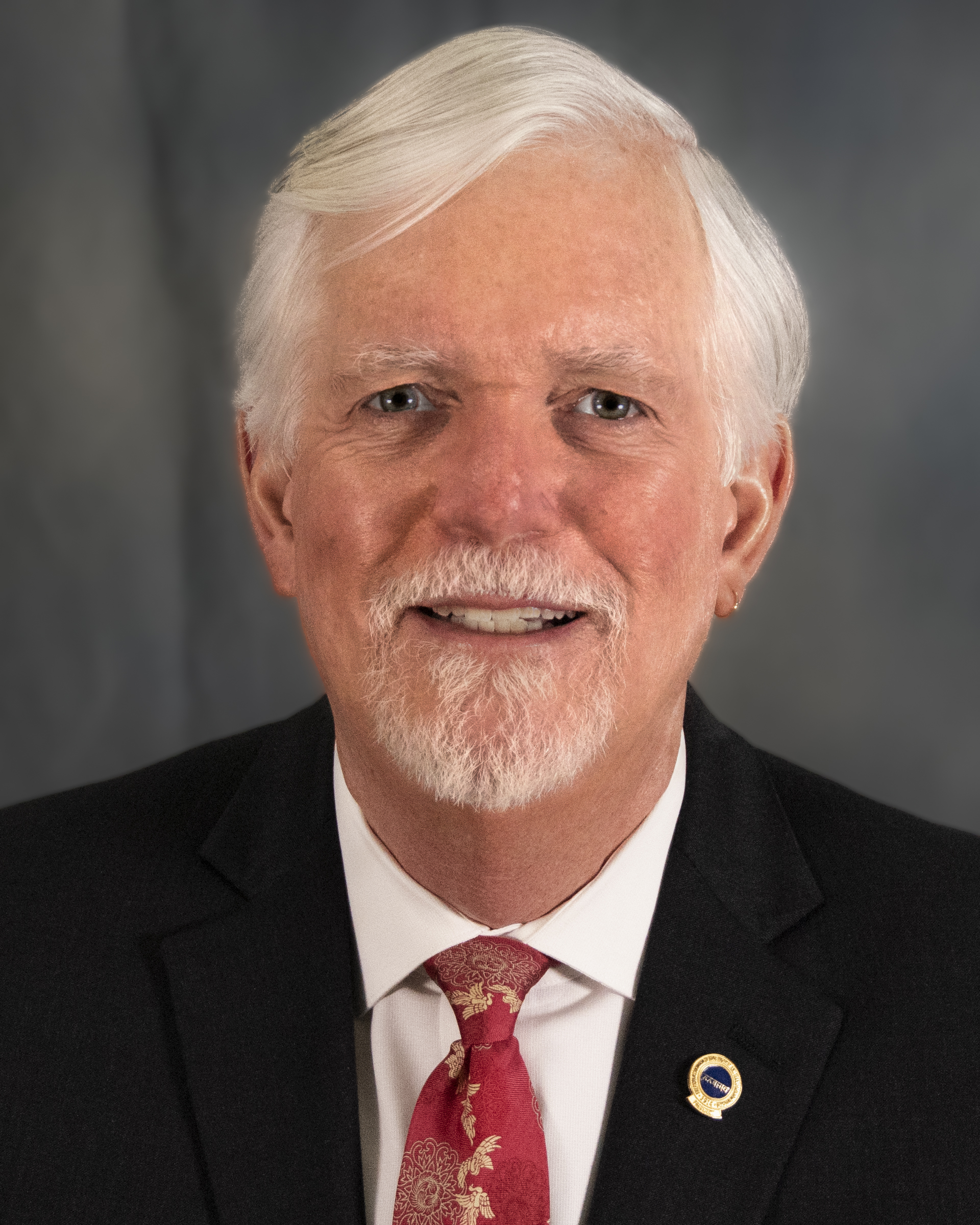 A smiling man with white hair, a white goatee, a small gold earring in his left ear, black suit, white shirt, lapel pin, and red and gold floral tie