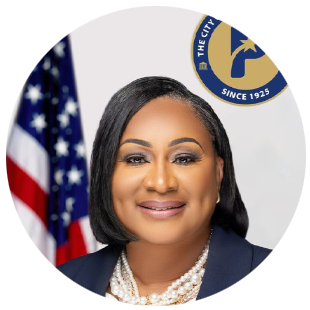 Speaker wearing a dark blazer and pearl necklace, posed against a white background with part of a U.S. flag visible