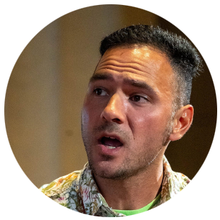 Speaker wearing a patterned shirt, shown speaking indoors against a softly lit neutral background.