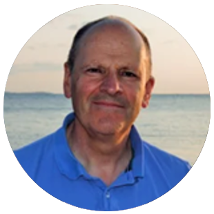Professional wearing a blue collared shirt, standing outdoors near a body of water with a distant shoreline in the background.