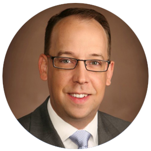 Professional wearing glasses, a dark suit jacket, and a light shirt with a tie, positioned against a brown studio background.