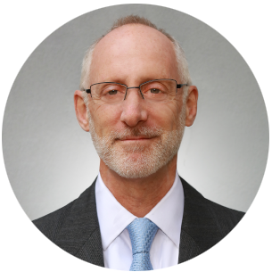 Professional headshot of a person wearing a dark suit jacket, white collared shirt, and light blue tie against a neutral gray background