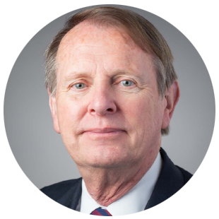 Professional headshot of a person wearing a dark suit jacket, white collared shirt, and red and blue striped tie against a neutral gray background