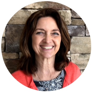 Speaker Pam Kenel wearing a coral cardigan over a patterned top, positioned in front of a stacked stone wall background.