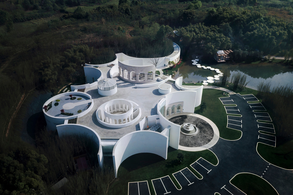 aerial view of a white pavilion with curving walls and a white stone path through the water to the trees beyond